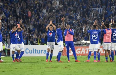 Image displaying footballers from Millonarios FC during a match. The photo shows the team in a blue kit clapping for the fans.