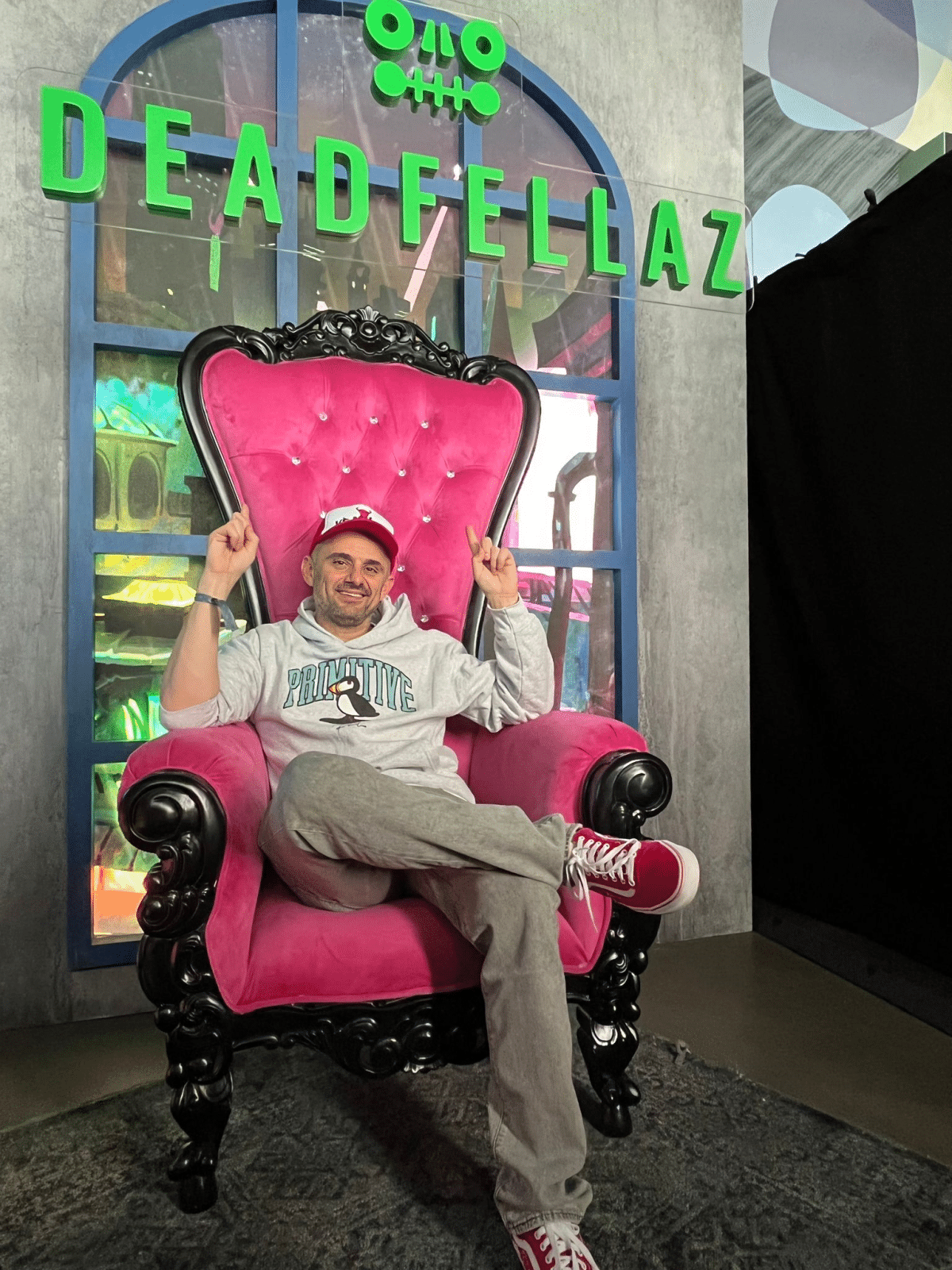 The picture shows Gary Vee sitting on a chair and pointing to a sign above his head that says DeadFellaz