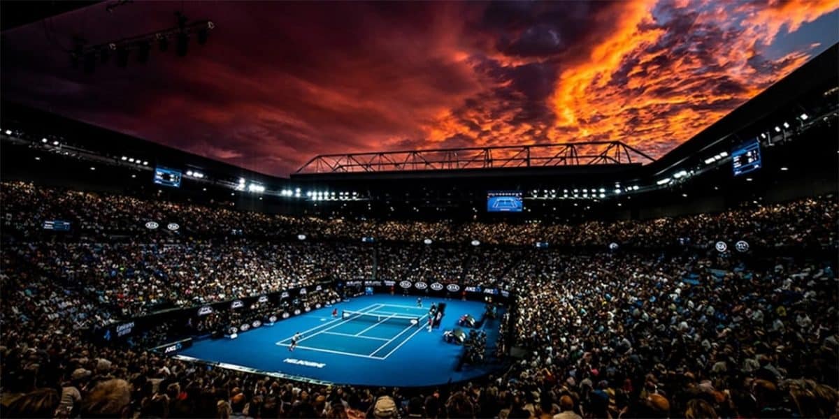 Una cancha de tenis azul está rodeada por un estadio lleno de gente, de noche con un cielo poniente.  Es el Abierto de Australia, que está profundizando en los beneficios de su programa NFT.