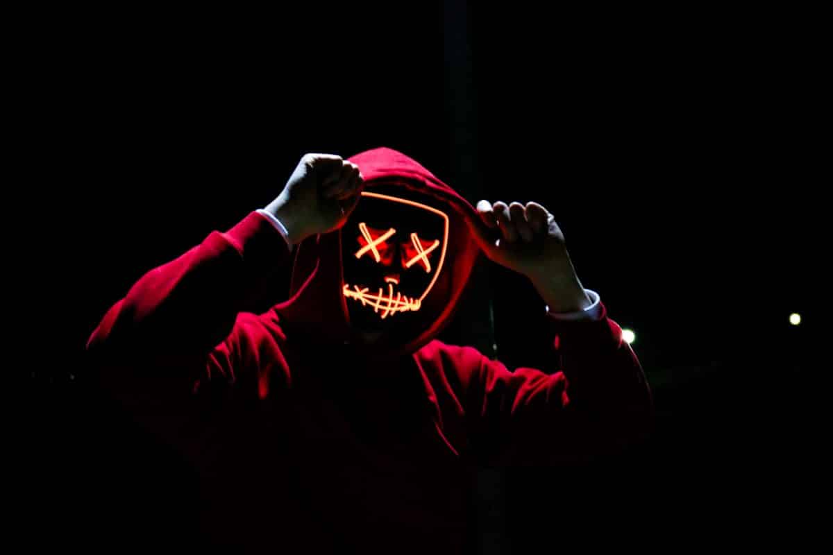 A man in a red sweatshirt holds the hood over his face, which is covered by a neon mask. Representative of the anonymous nft thief monkey drainer