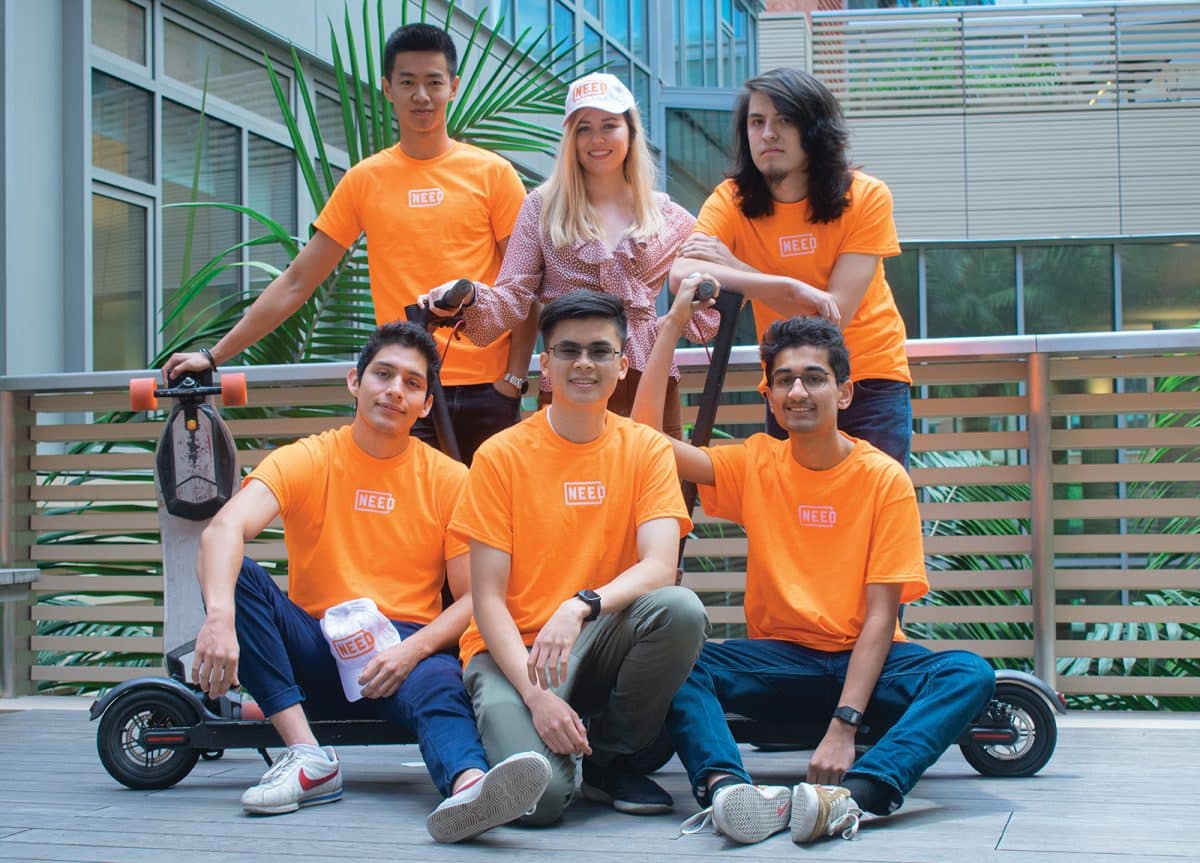 Seis estudiantes universitarios con camisas de color naranja brillante se sientan y se paran frente a un scooter eléctrico.  Uno de ellos es Frank DeGods, que acaba de drogarse.