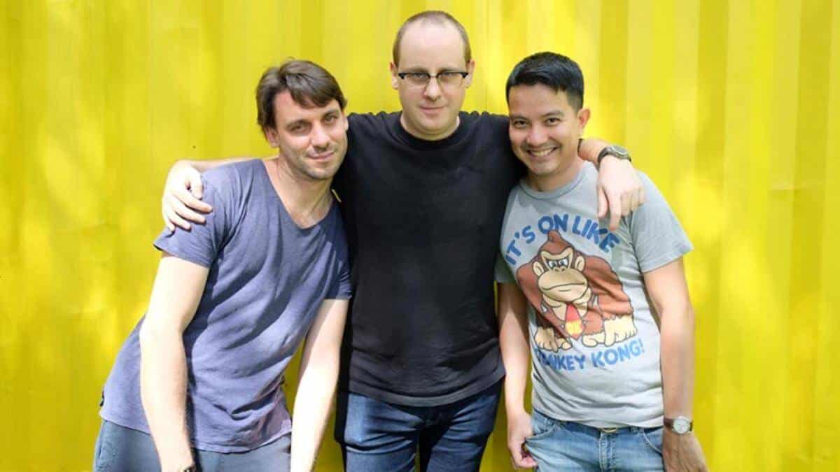 Three men in t-shirts stand before a yellow background in support of Mighty Bear Games releasing Mighty Action Heroes.