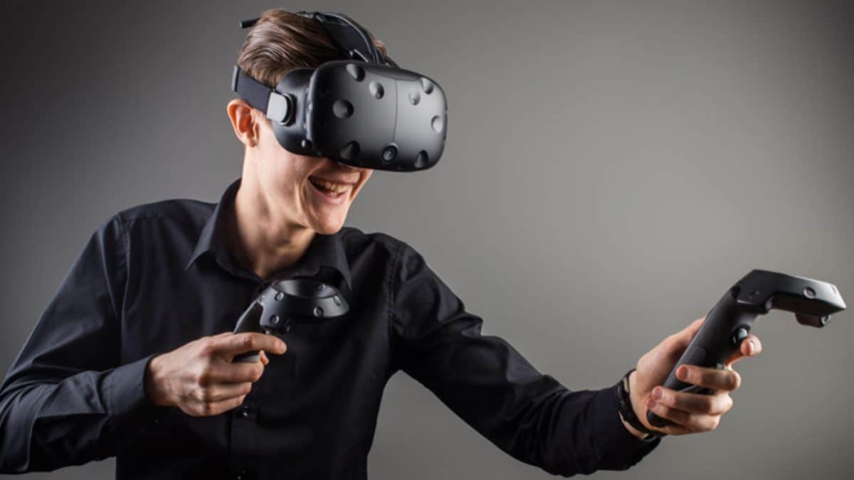 A man stands before a gray backbground with a VR headset and controllers on.
