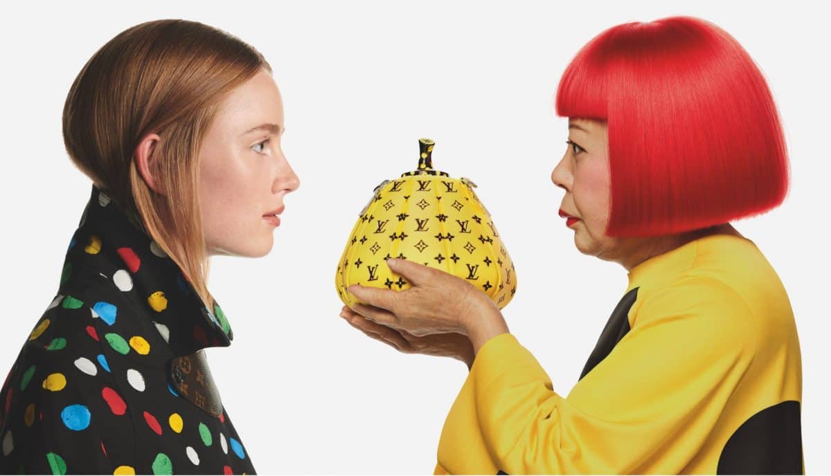 Yayoi Kusama holds a yellow pumpkin with Louis Vuitton emblems in front of a model in support of the Louis Vuitton Yayoi Kusama NFT collection.