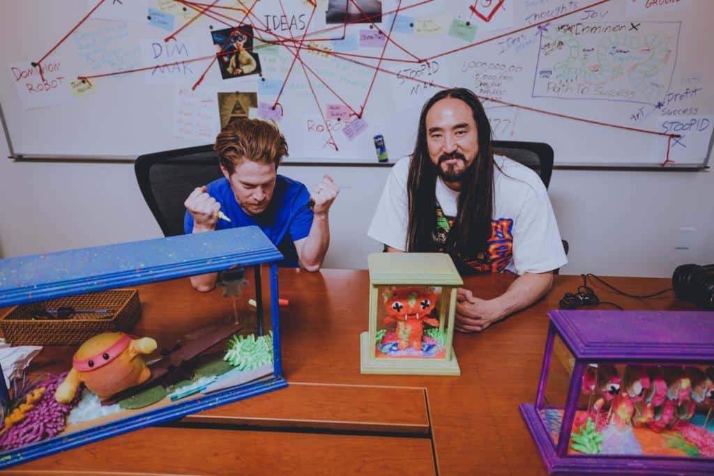 a picture of Steve Aoki & Seth Green, sitting at a desk with a physical avatar copy of Aoki's Replicant X NFT project.