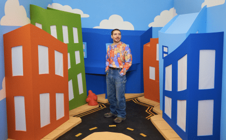 The artist, Danny Cole, stands in front of cartoon buildings while wearing a shirt with artwork on it.