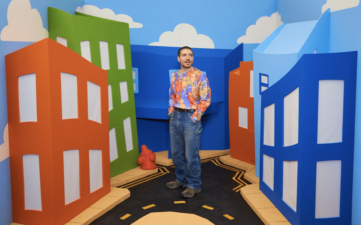Artist Danny Cole wears an artwork shirt and stands in front of cardboard buildings.